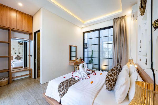 Romantic Balinese bedroom with king bed adorned in batik linens, swan towel, rose petals, and balcony view through large glass doors.
